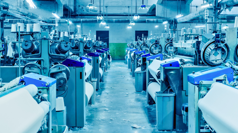 Rows of industrial textile looms with large spools of white thread, illuminated by overhead lights in a factory setting.