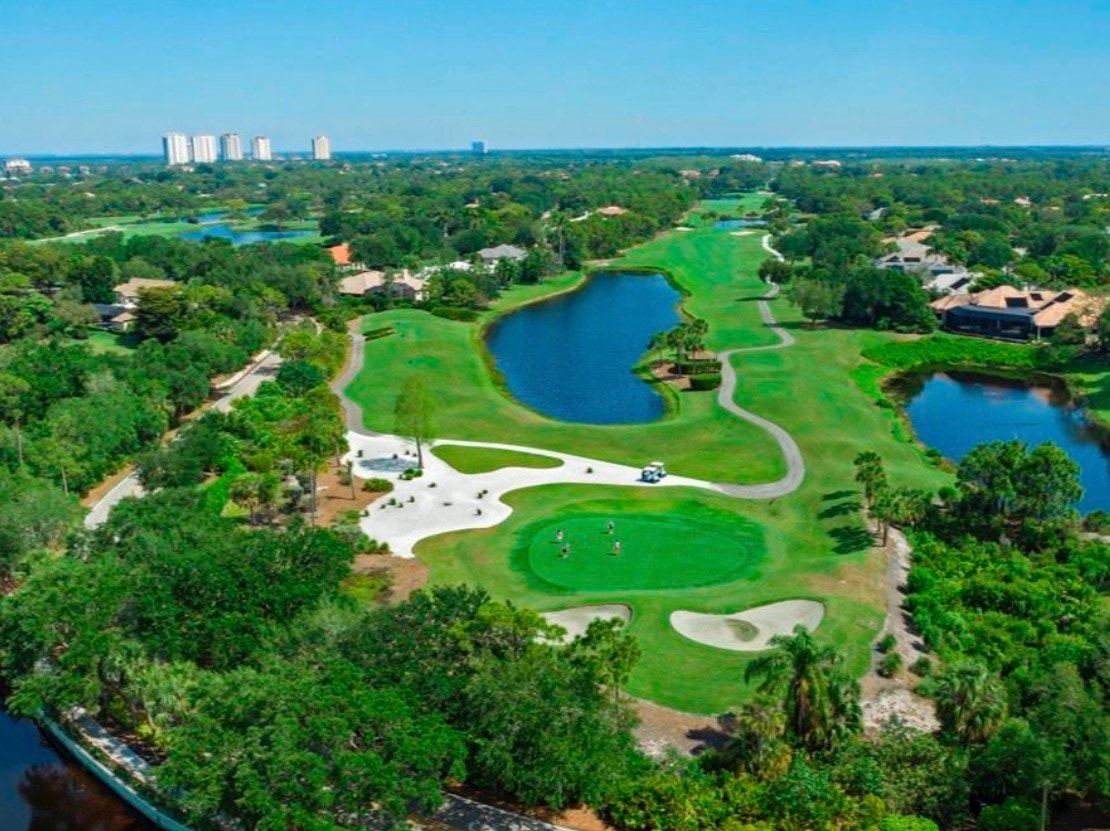 The image depicts a luxurious single-family home showcasing Spanish-Revival architecture, surrounded by lush mature landscaping that enhances its elegance. This exquisite residence is located in the amenity-rich Pelican Landing community, known for its prestigious golf courses and beautiful tropical environment.
