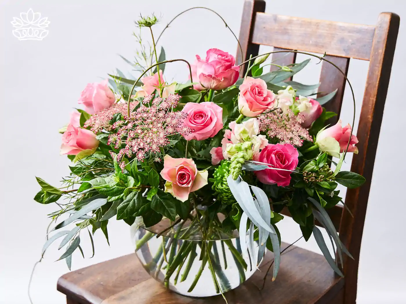 Elegant pink rose bouquet with lush greenery and soft pink accents, artfully arranged in a clear vase on a vintage wooden chair, ideal for front desk decor or reception desk.