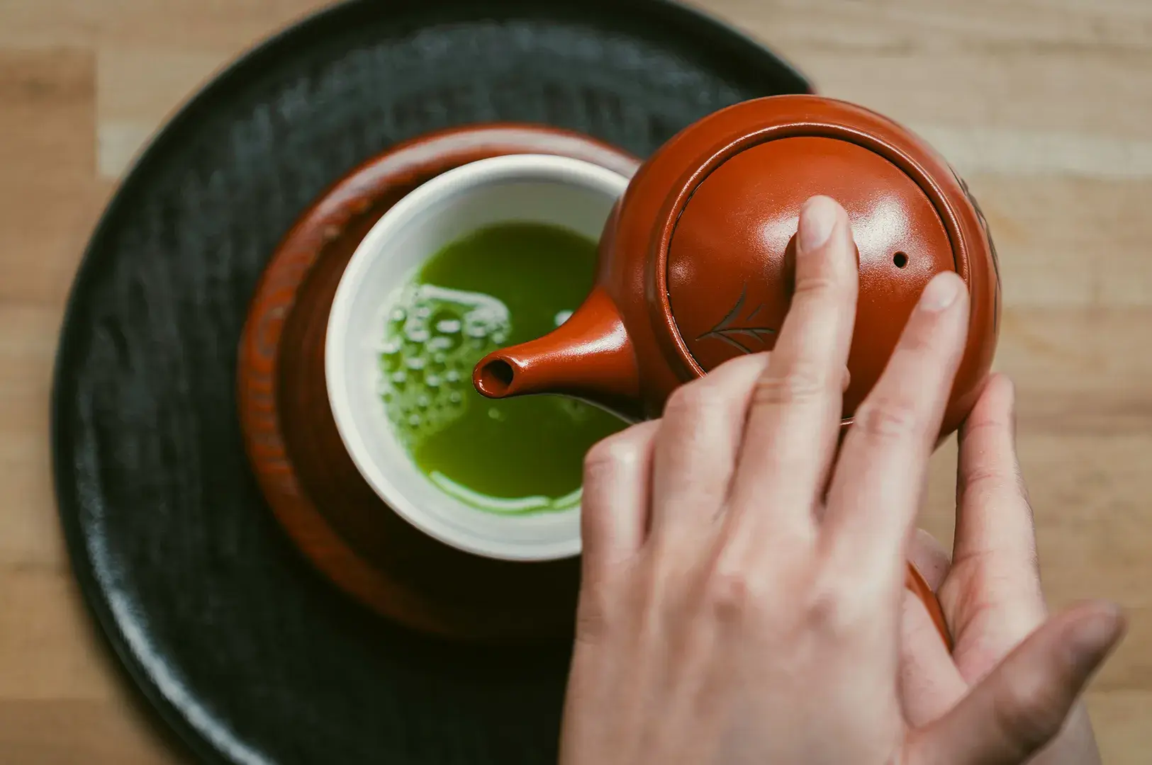Person pouring vibrant green tea from a traditional reddish-brown ceramic teapot into a small white cup resting on a black tray.