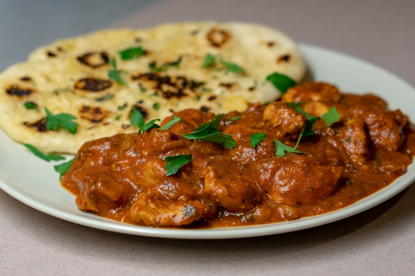 Plate with creamy butter chicken garnished with herbs, accompanied by charred naan bread. The dish appears rich and flavorful.