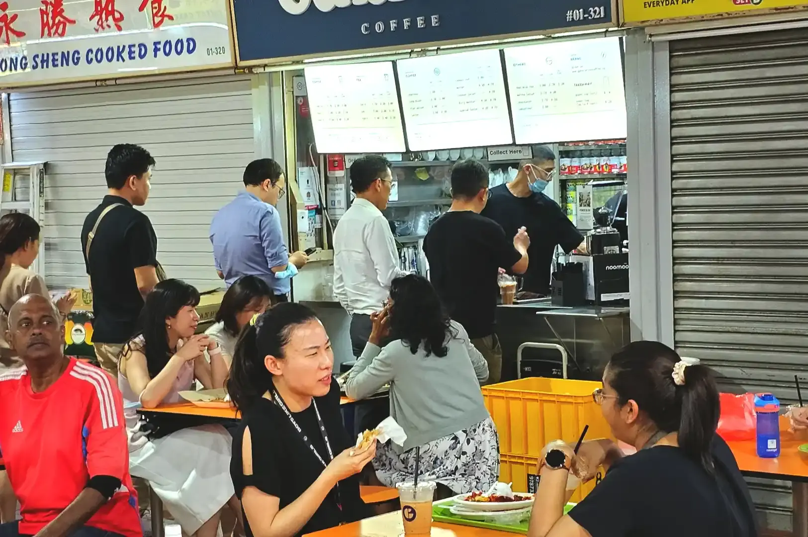 A bustling dining scene with customers seated at orange tables outside Generation Coffee, enjoying drinks and meals at Tekka Centre.