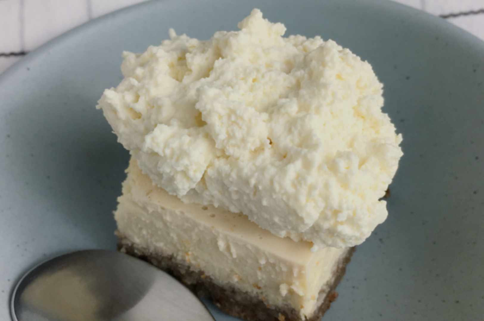 A slice of cheesecake with a crumbly base, topped with a thick layer of whipped cream on a light blue plate. A silver spoon is placed beside it.