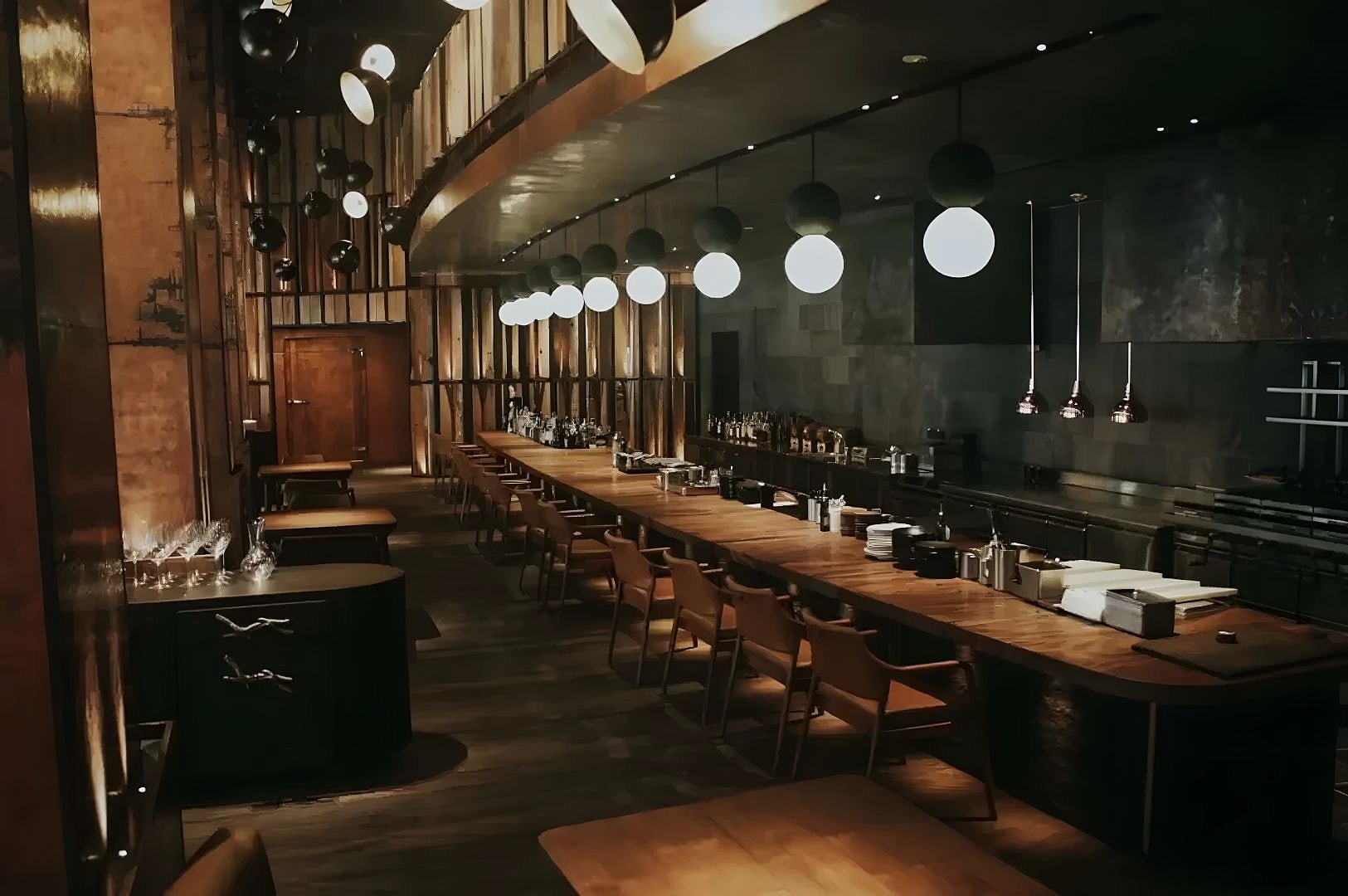 A warm, inviting restaurant interior with a long wooden bar, surrounded by chairs. Soft, round hanging lights create an elegant, cozy atmosphere.