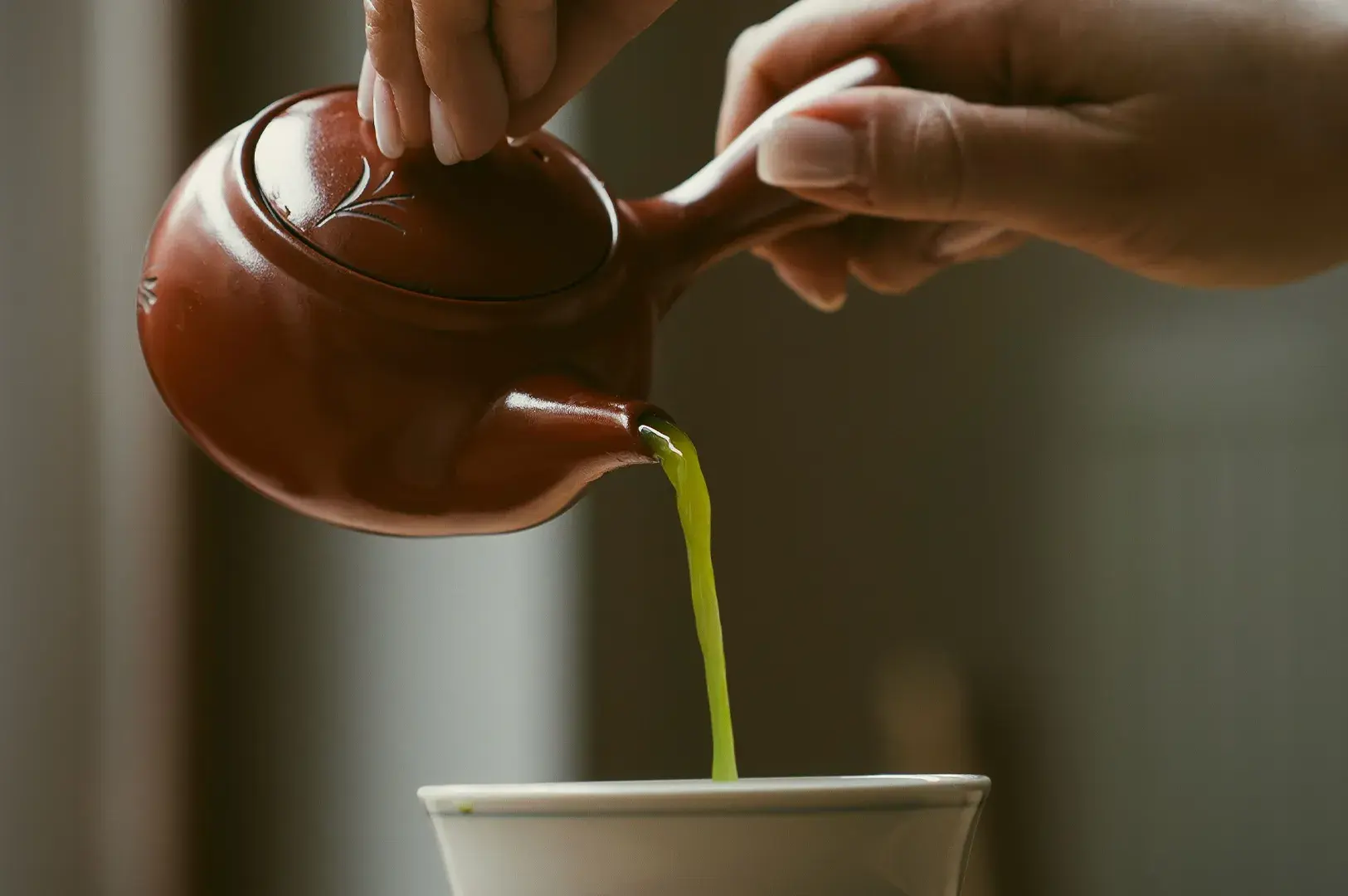Close-up shot of a person pouring bright green tea from a reddish-brown ceramic teapot into a white cup.
