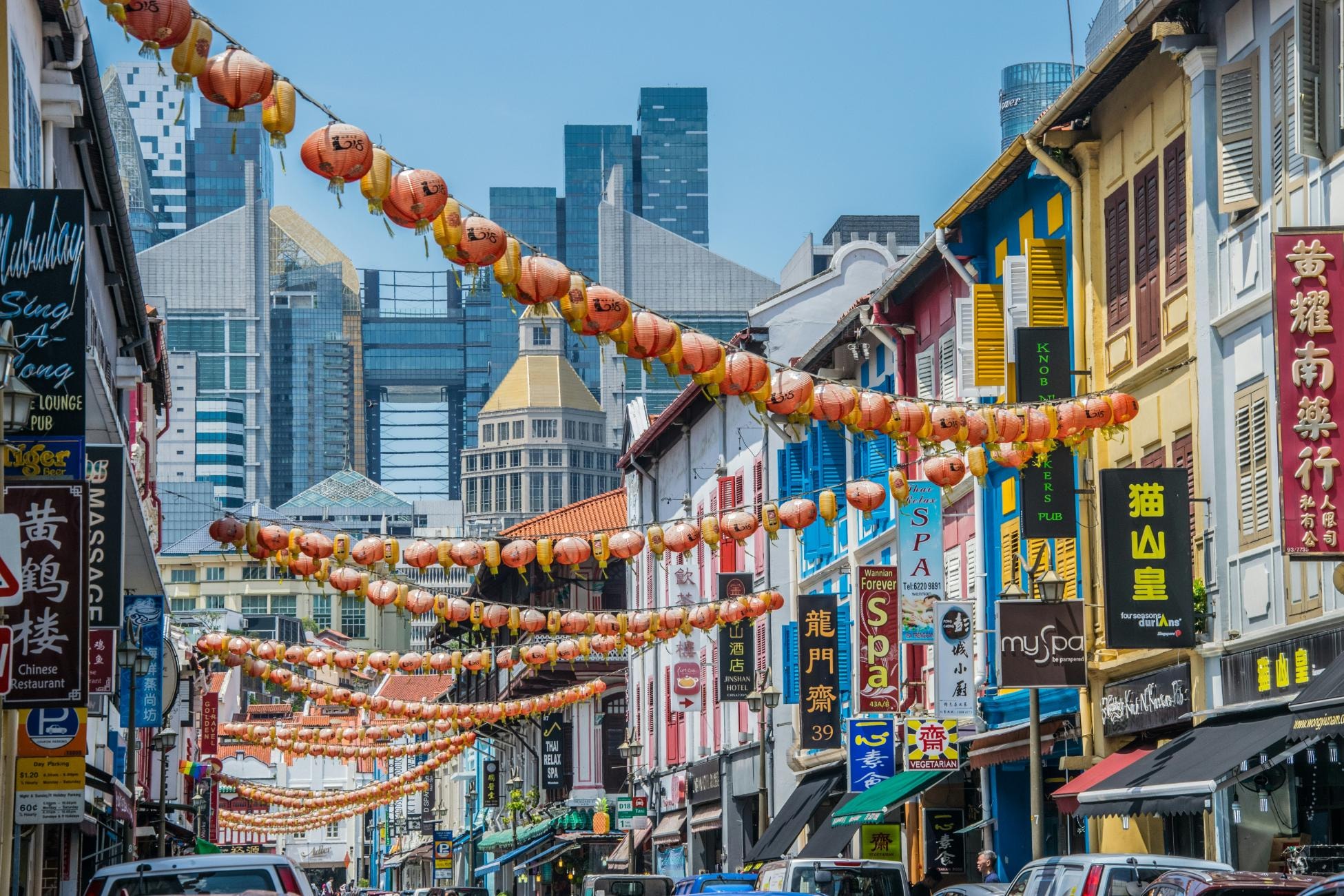 This vibrant street scene features rows of colorful traditional shophouses adorned with strings of red lanterns overhead, creating a festive atmosphere in the bustling district. Behind the historic two-story structures, a modern skyline of glass skyscrapers and a golden-domed tower rises steeply, creating a striking contrast between the city's heritage and its contemporary development.