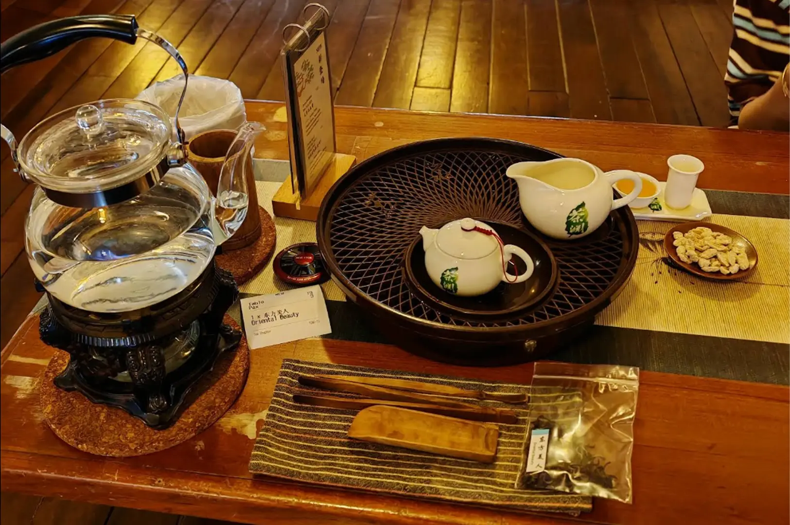 A high-angle, eye-level shot of a tea service on a wooden table. A glass teapot sits on a warmer to the left. In the center, a dark, circular tea tray holds a small white teapot and a matching pitcher with green tea-leaf logos. Next to the tray, there is a small cup of tea, a bowl of snacks, and a sealed packet of "Oriental Beauty" tea. Wooden tea tools lie on a striped cloth in the foreground.