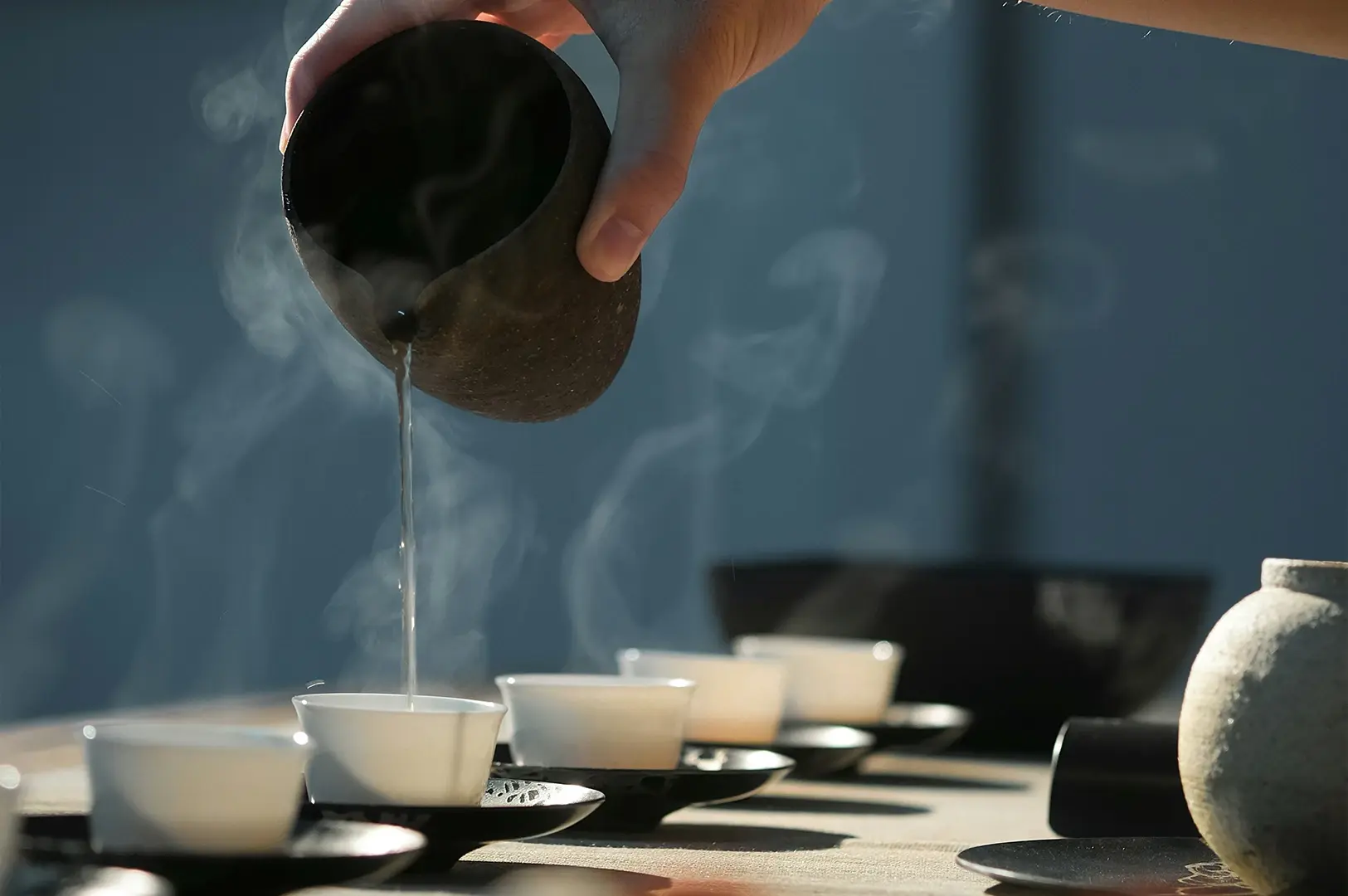 A close-up shot of tea being poured from a dark, rustic clay vessel into a row of white tea cups. Steam billows upward from the hot liquid, partially obscuring the background. The scene is brightly lit, emphasizing the movement of the liquid and the delicate ceramic textures.