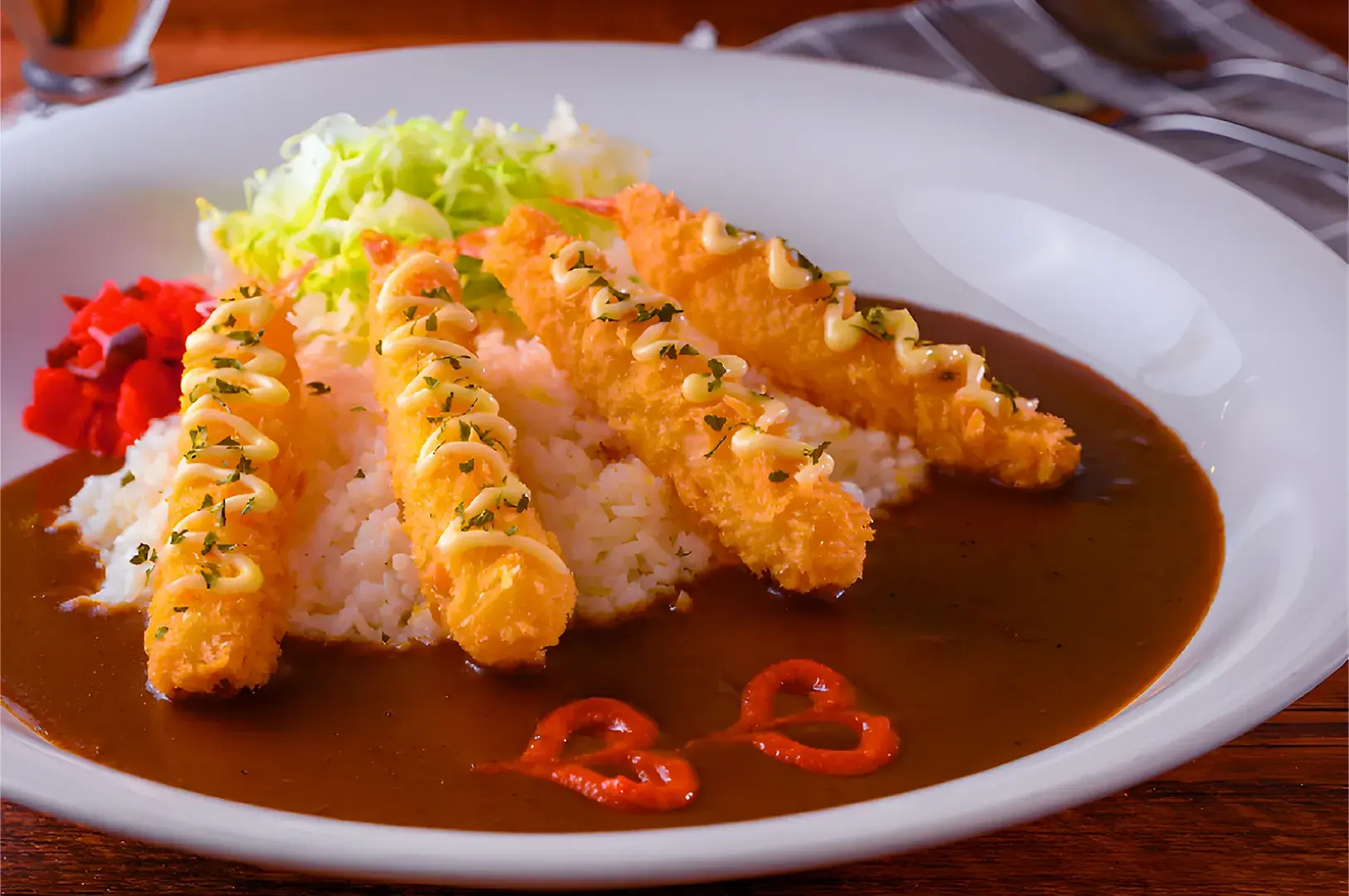 Low-angle close-up shot of Japanese pork katsu curry served on a white plate, featuring sliced golden-breaded tonkatsu over rice topped with rich brown curry sauce, shrimp tempura, shredded cabbage, and pickled vegetables on a wooden table.