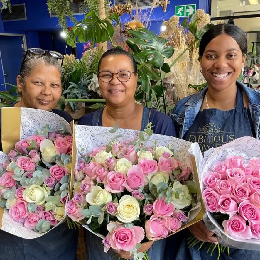 Three smiling Fabulous Flowers team members holding beautifully wrapped bouquets of pink and white roses with green foliage, standing in a lush and vibrant flower shop.