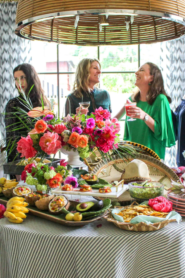 Guests chatting and holding cocktails beside a colorful buffet table decorated with vibrant floral arrangements, fruit, and appetizers in a bright indoor space.
