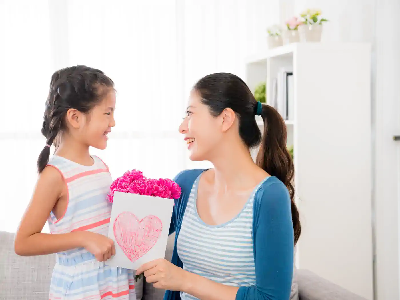 A joyful young girl presents her mother with a handmade card and a box of vibrant pink flowers, celebrating a special moment together. Just Because Flowers. Delivered with Heart. Fabulous Flowers and Gifts