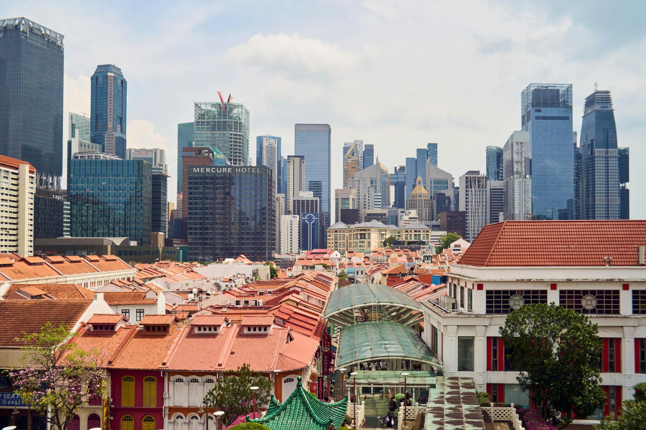 This low-angle shot captures the tiered, dark blue roof of a traditional Chinese structure topped with a golden finial, contrasted by a tall tree with brown foliage on the left. On the right, an ornate green-tiled roof ridge adorned with small mythical figurines juts into the frame, with a pale moon visible in the clear blue sky between the buildings.