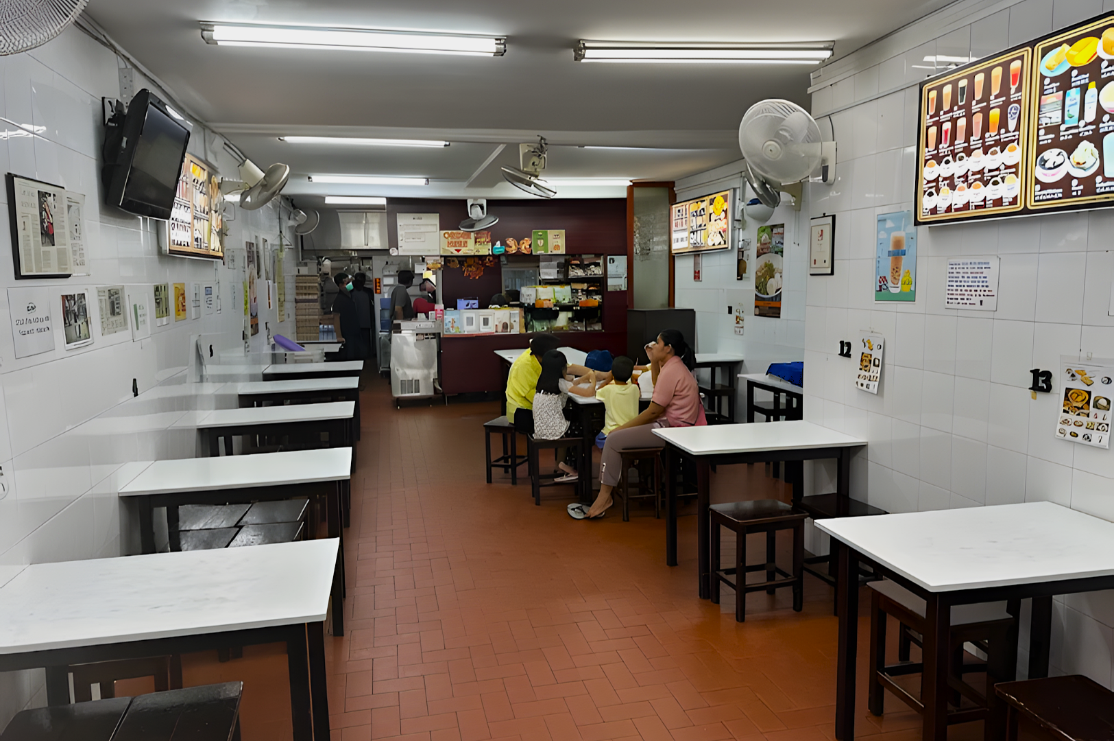A cozy, quiet restaurant with white tiled walls and red-brown flooring. A family sits at one of the wooden tables near a counter displaying menu options.
