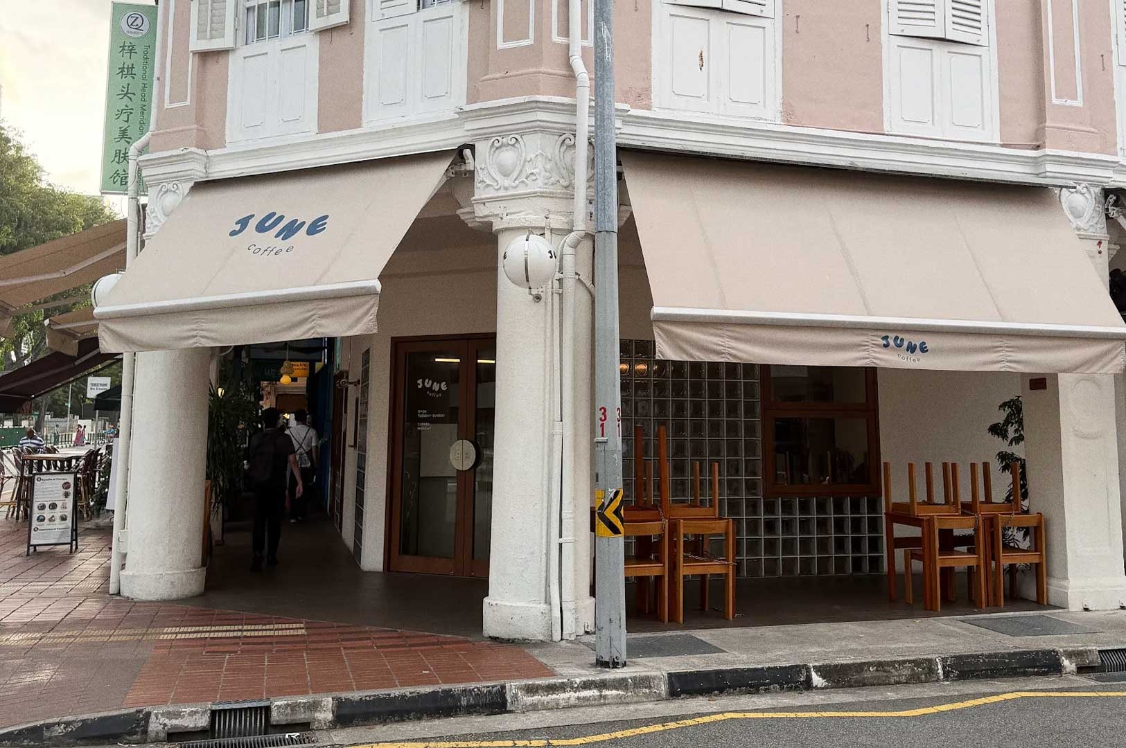 Street‑level view of June Coffee storefront on a tiled sidewalk, shuttered windows above, and stacked wooden seating outside the café.