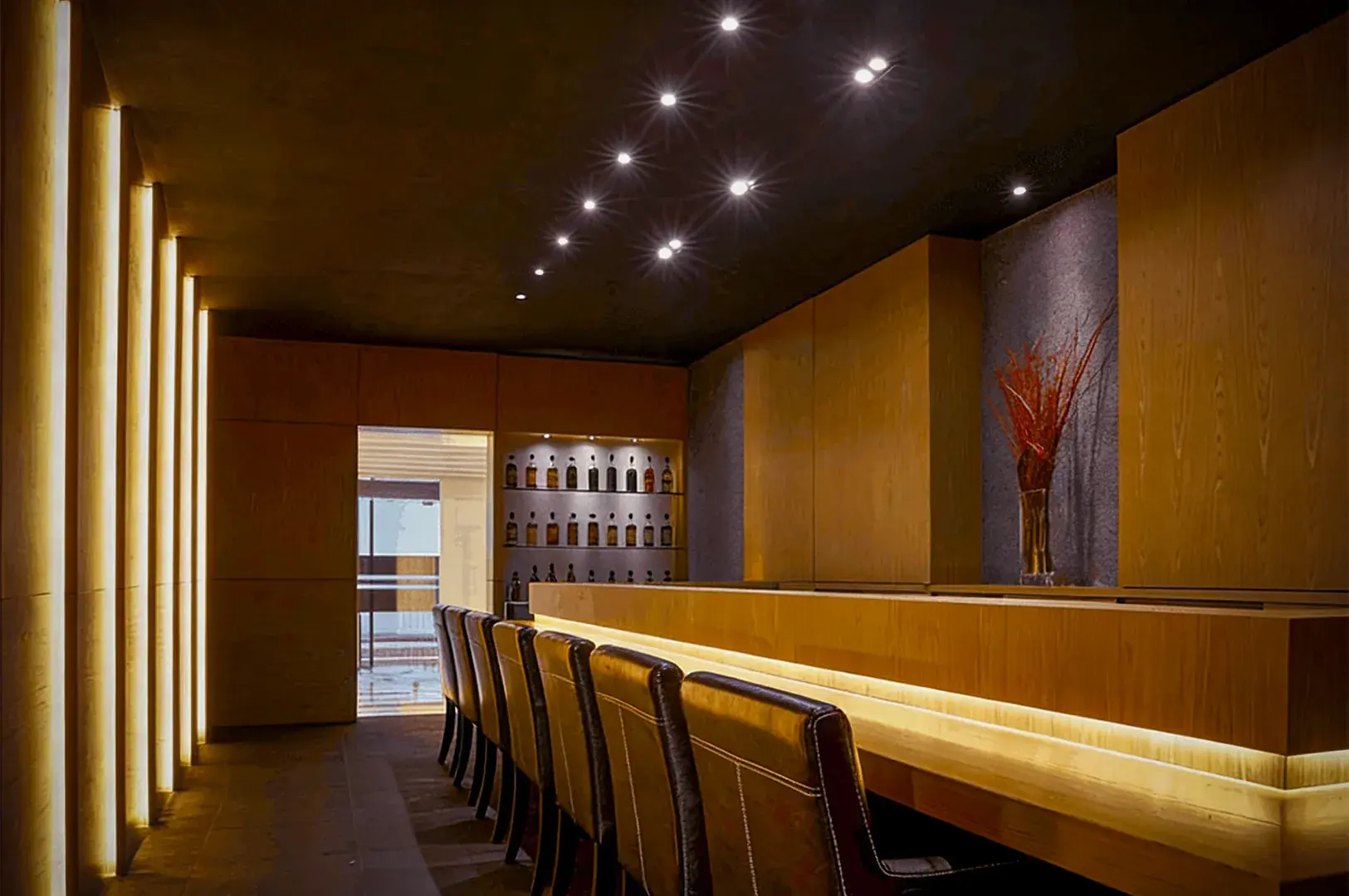 A modern bar with a wooden interior, featuring a row of leather chairs, a softly lit countertop, bottles on shelves, and dim overhead lighting.