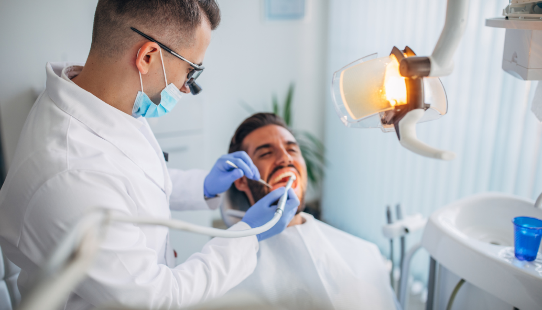Tannlege med munnbind undersøker munnen til en smilende pasient i tannlegestol under sterkt lys på tannlegekontor.