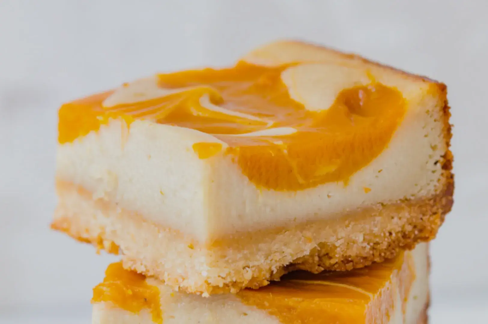 A close-up of a slice of cheesecake with a swirled mango topping on a graham cracker crust, stacked on top of another slice.