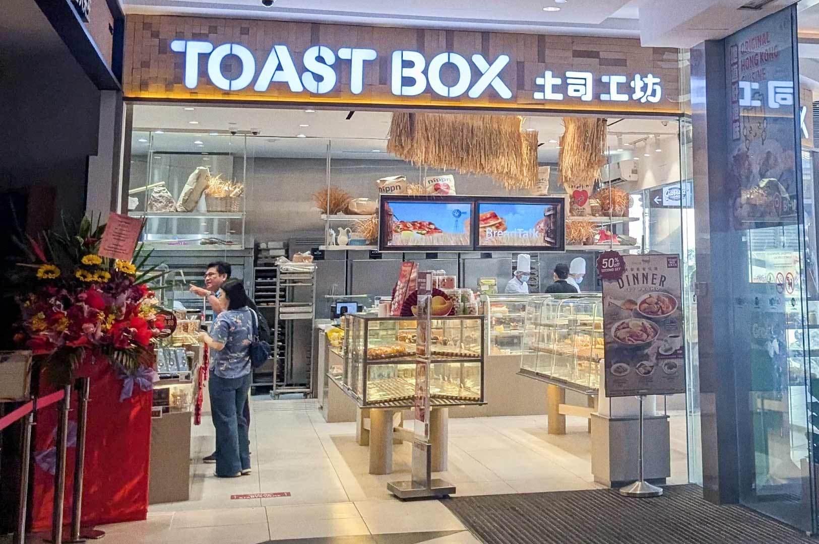 Mall storefront of Toast Box showing glass display cases filled with pastries, digital menu screens above the counter, staff serving customers, and a promotional stand advertising a dinner set.