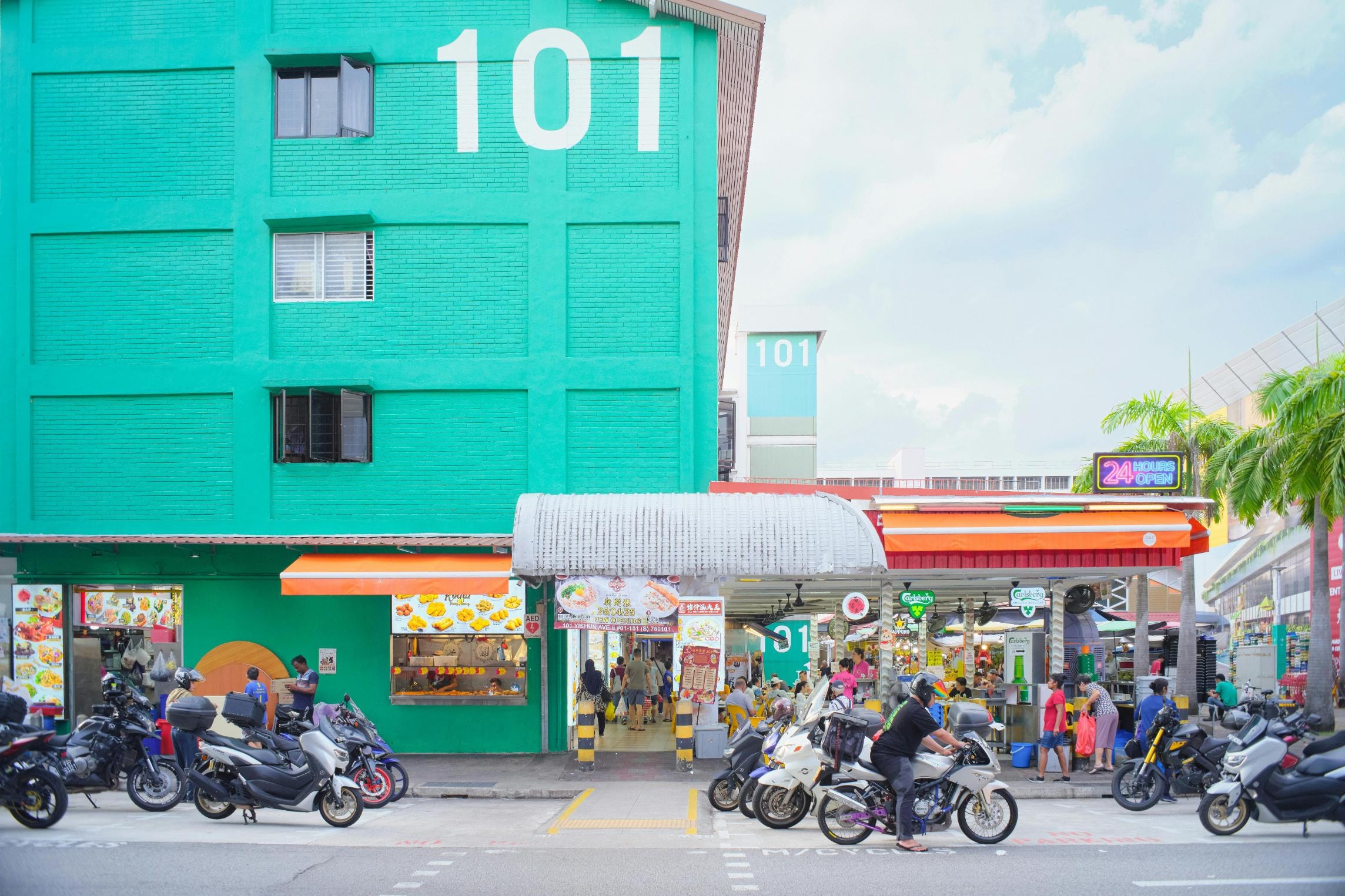 A vibrant turquoise building, prominently marked with a large white "101," rises above a bustling street scene lined with rows of parked motorcycles. Beneath the residential block, a lively open-air eatery features colorful food stalls and orange awnings, welcoming diners with a bright "24 Hours Open" neon sign.