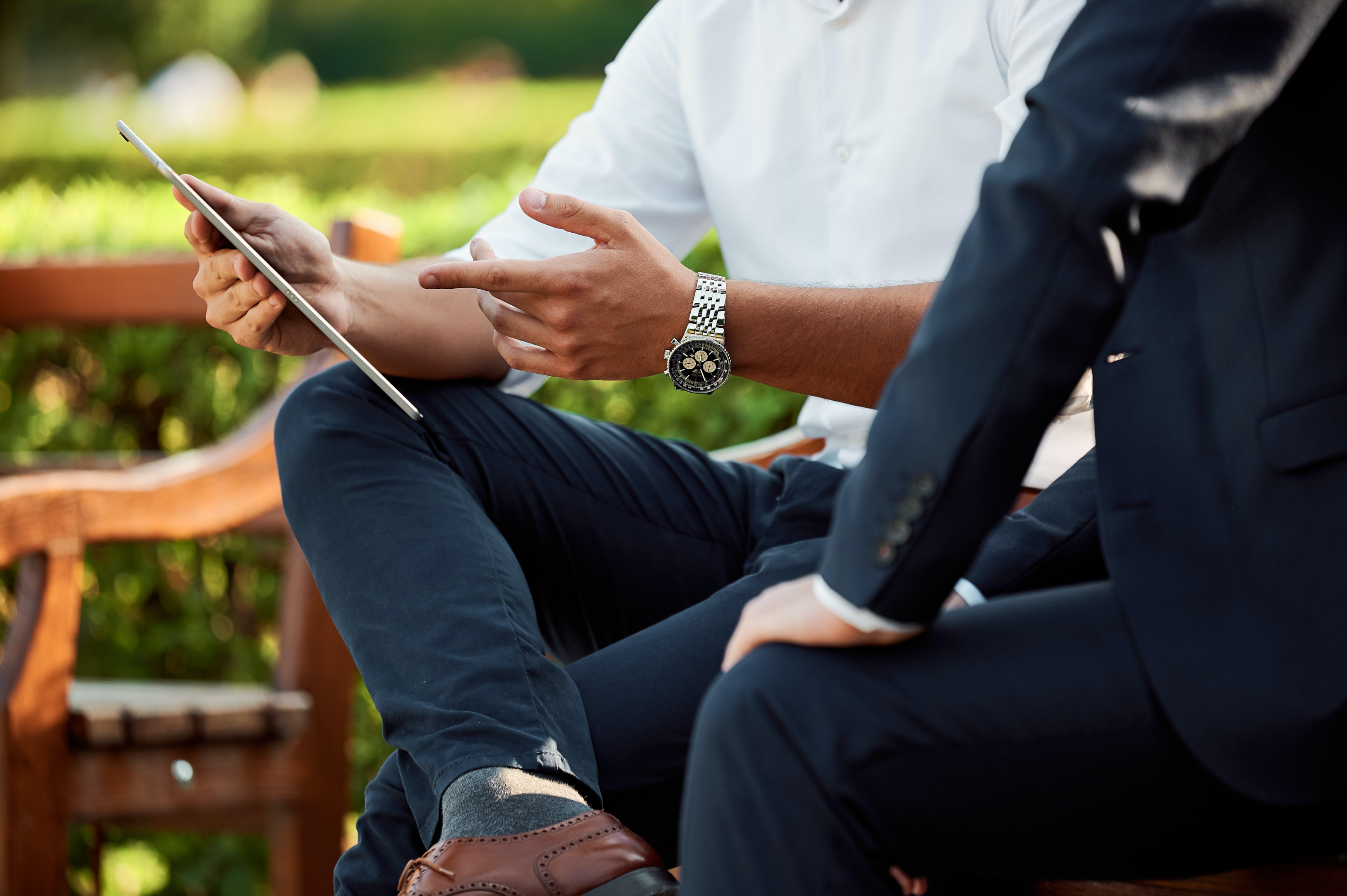 Photo of two people sitting on a bench during the day. Shot is focused on the person's hand holding an iPad.