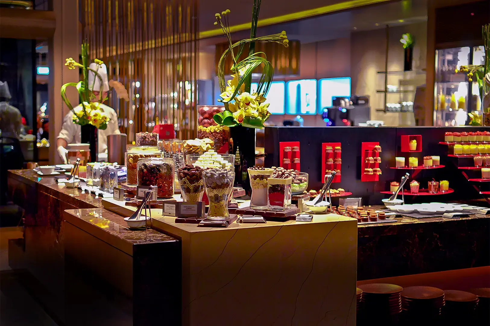 Luxurious buffet with a variety of desserts in jars, surrounded by elegant flower arrangements in a warmly lit setting, creating a sophisticated ambiance.