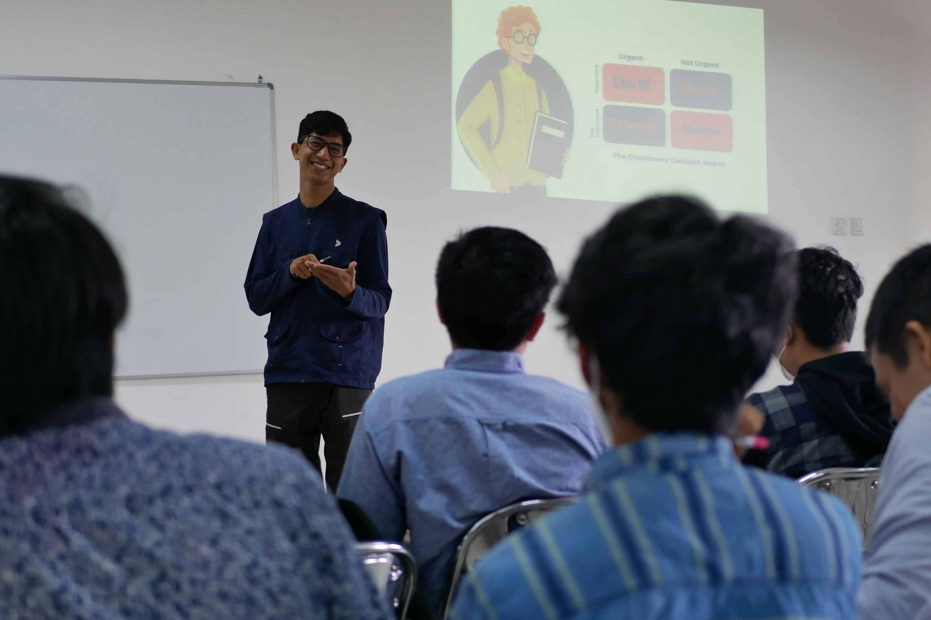This image captures a smiling speaker presenting a lecture to an attentive audience in a classroom setting. Behind him, a projector screen displays a slide about the "Eisenhower Decision Matrix," suggesting a workshop focused on productivity and time management.
