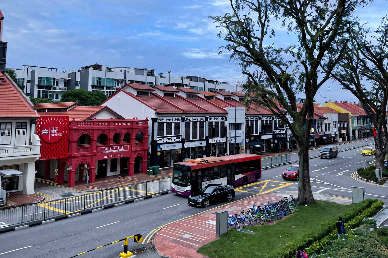 Street view of a row of restored shophouses in various colors, including a prominent red building with arched windows. A public bus and several cars travel along the road, while bicycles are parked at a docking station. Tall trees line the street, with modern low-rise apartments visible in the background under a cloudy sky