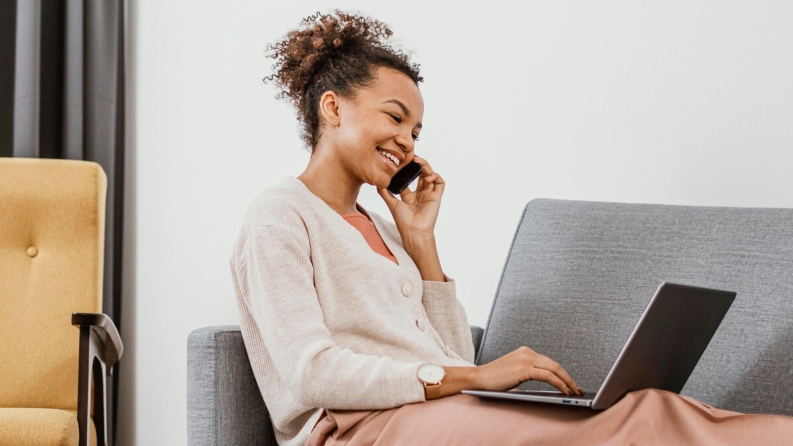 Smiling woman working remotely from her living room, speaking on a mobile phone while using a laptop, representing how cloud-based business VoIP solutions enable seamless communication for hybrid and remote teams.