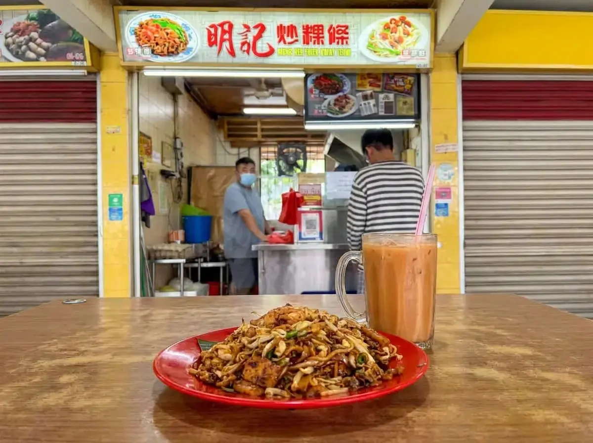 "A red plate of stir-fried Char Kway Teow noodles and a glass mug of iced milk tea on a wooden table, in front of the 'Meng Kee Fried Kway Teow' hawker stall."