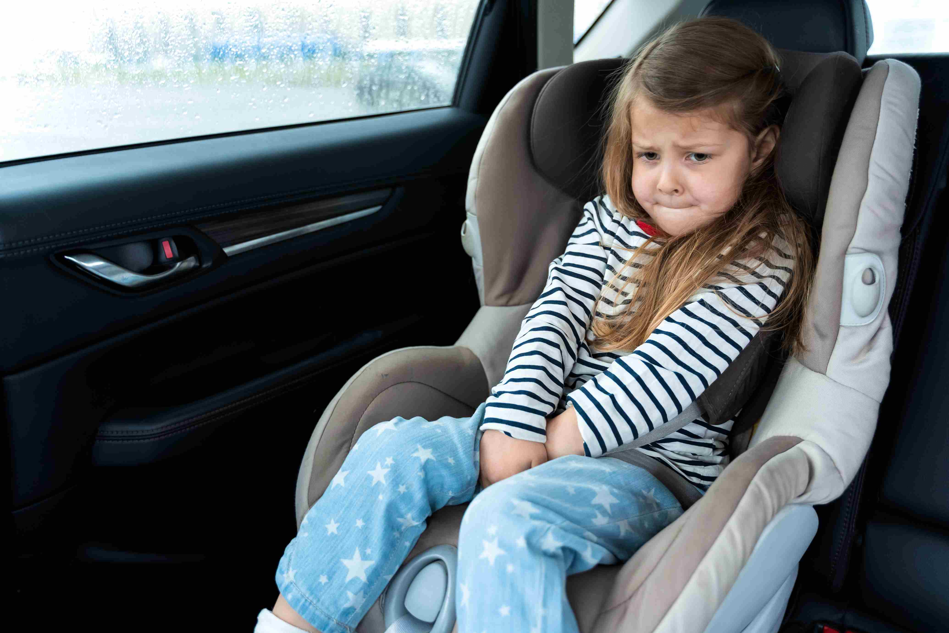 Mädchen im Kindersitz im Auto hält sich verkrampft und verzieht das Gesicht, weil sie dringend aufs Klo muss 