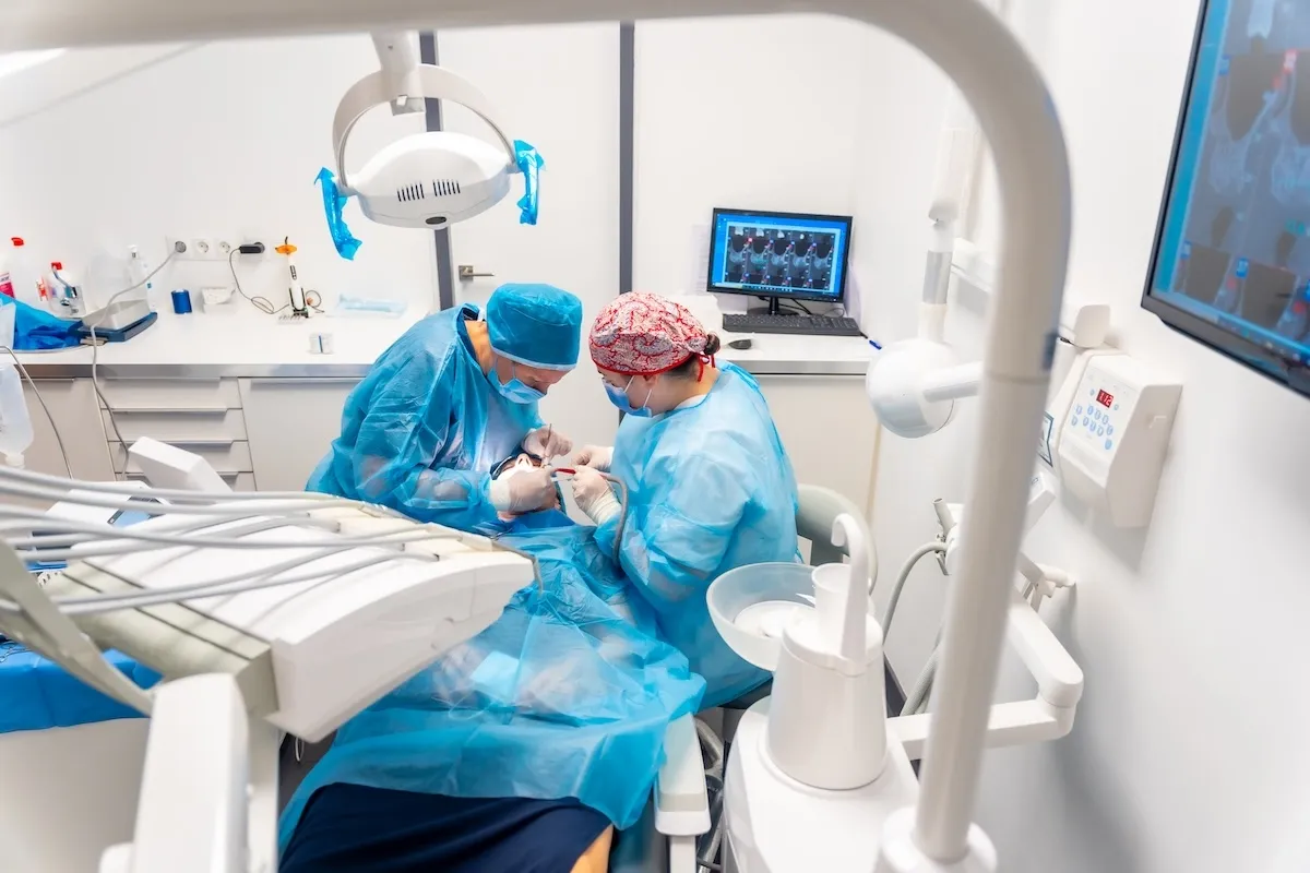Dental surgery procedure being performed by two surgeons in sterile blue gowns and caps with advanced dental equipment and digital monitoring technology, as part of the dental practice.