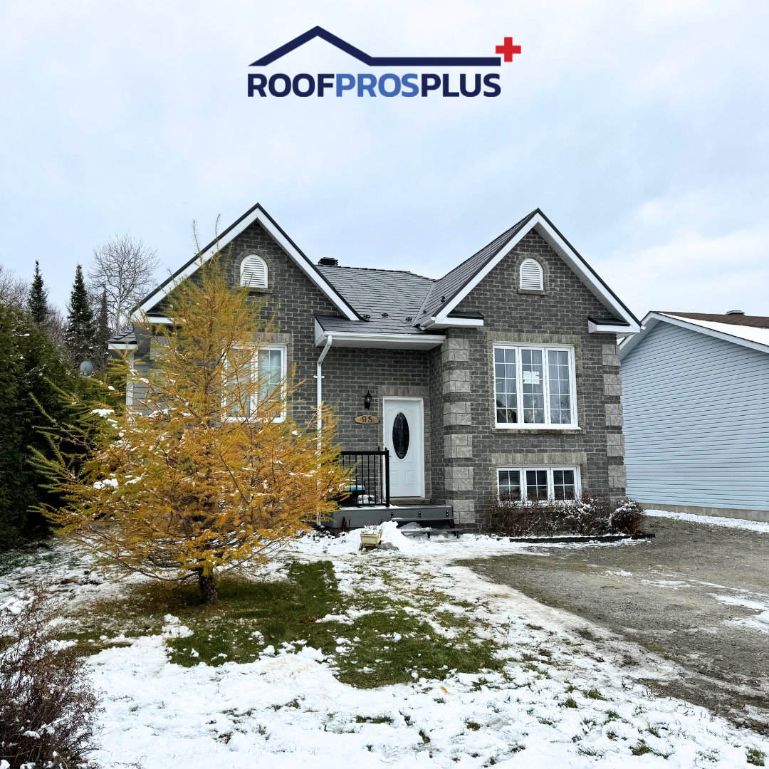 A grey brick house with a pointed roof, large front windows, and white trim sits against a cloudy sky. A snow-dusted lawn with a small yellow-leaved tree is in front of the house. The logo for RoofProsPlus is at the top of the photo. 