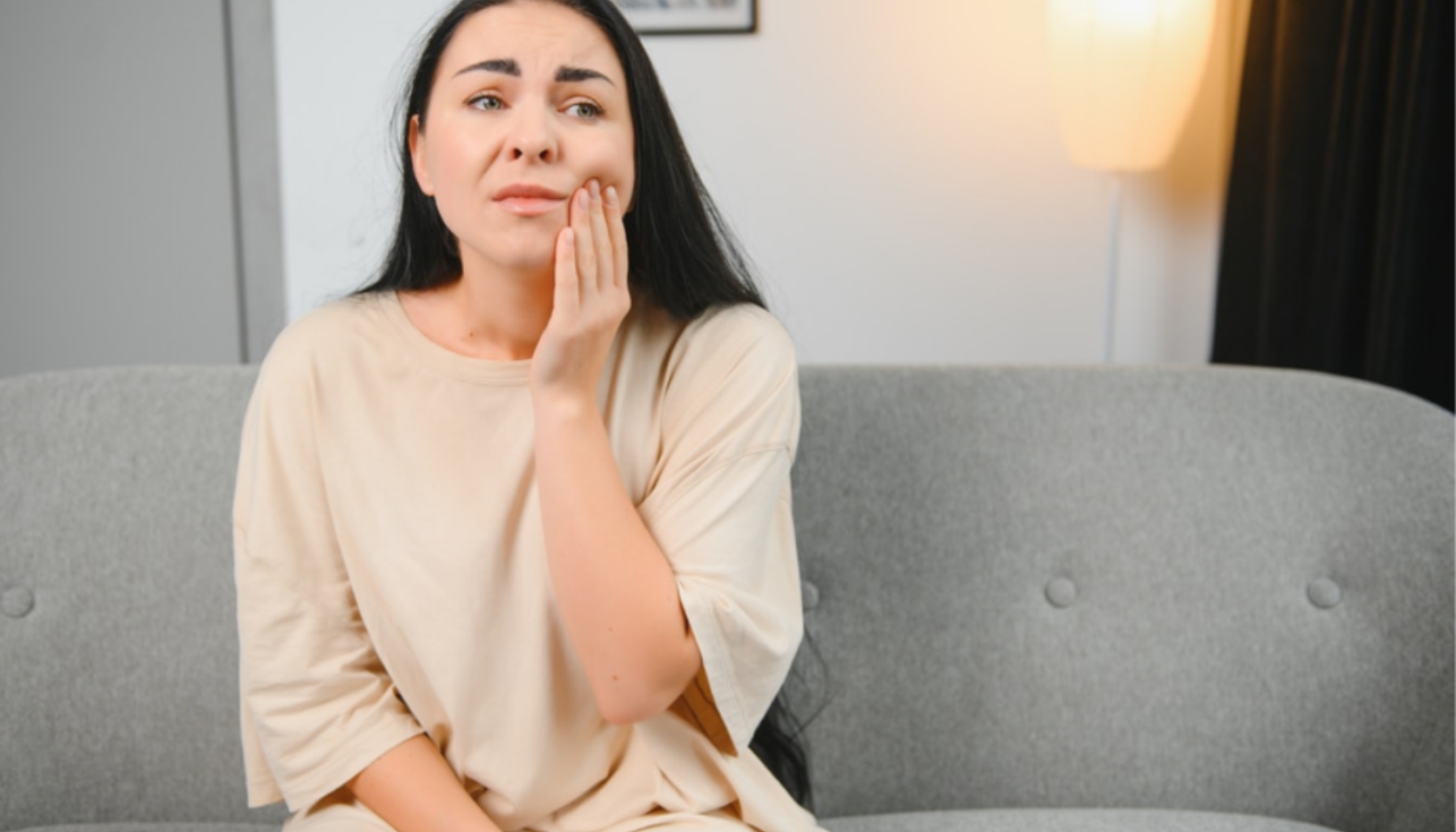 Betennelse Etter Trekking av Tann: Når Helingen Går Galt 15 Kvinne sitter på sofa hjemme og holder hånden mot kinnet, med tydelig uttrykk for tannverk eller smerte ansiktet.