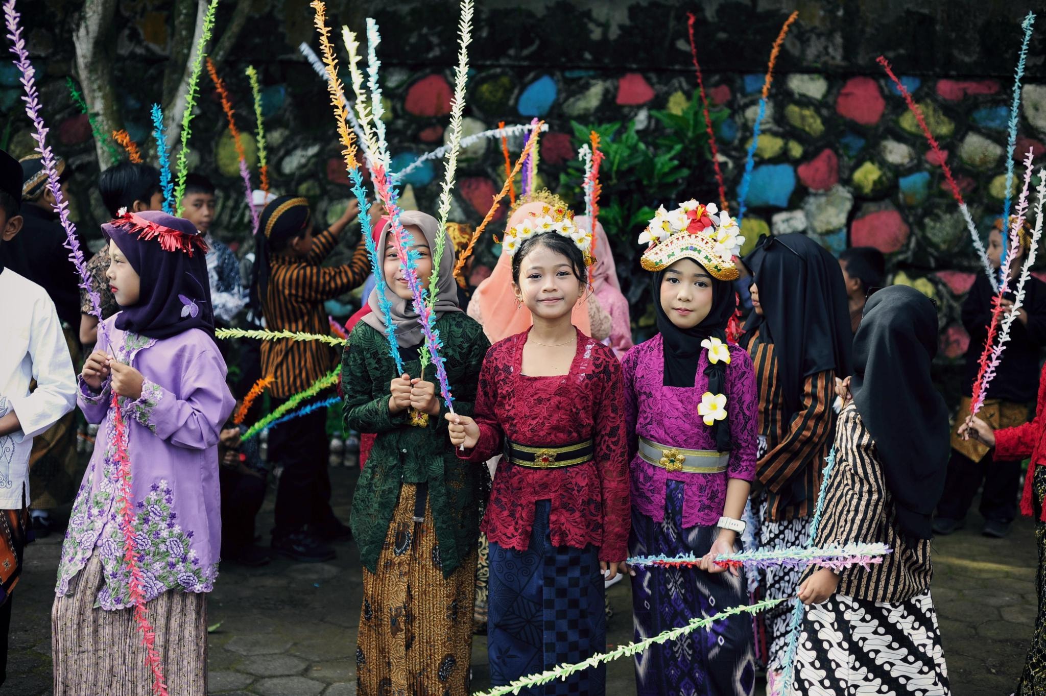 This image captures a group of children dressed in vibrant traditional attire, with girls wearing colorful kebayas, sarongs, and ornate floral headdresses. They are holding slender, multi-colored decorative sticks while standing before a textured stone wall, creating a scene that conveys a festive and culturally rich atmosphere.
