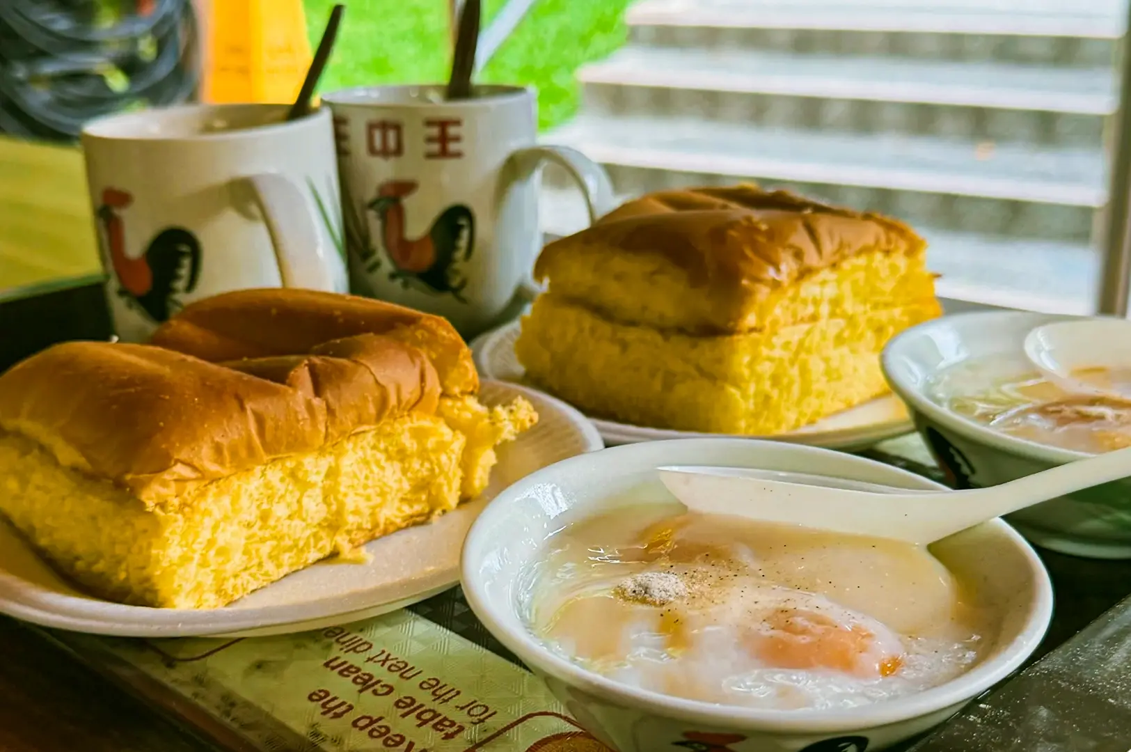 Close‑up eye‑level shot of a traditional Singapore breakfast featuring kaya toast on white plates, soft‑boiled eggs in bowls, and coffee in ceramic mugs with rooster designs, placed by a window with natural daylight.