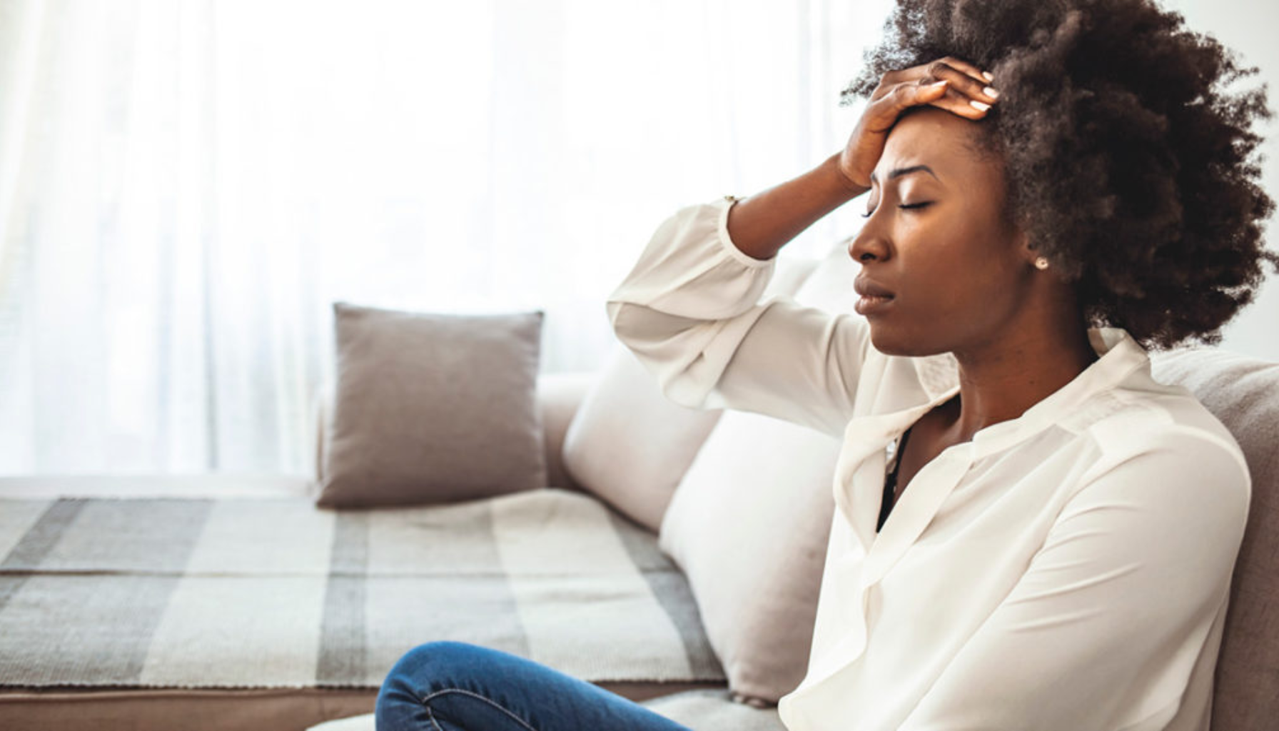 Kvinne sitter på sofa med hånden mot pannen og ser tydelig stresset ut i et lyst rom.