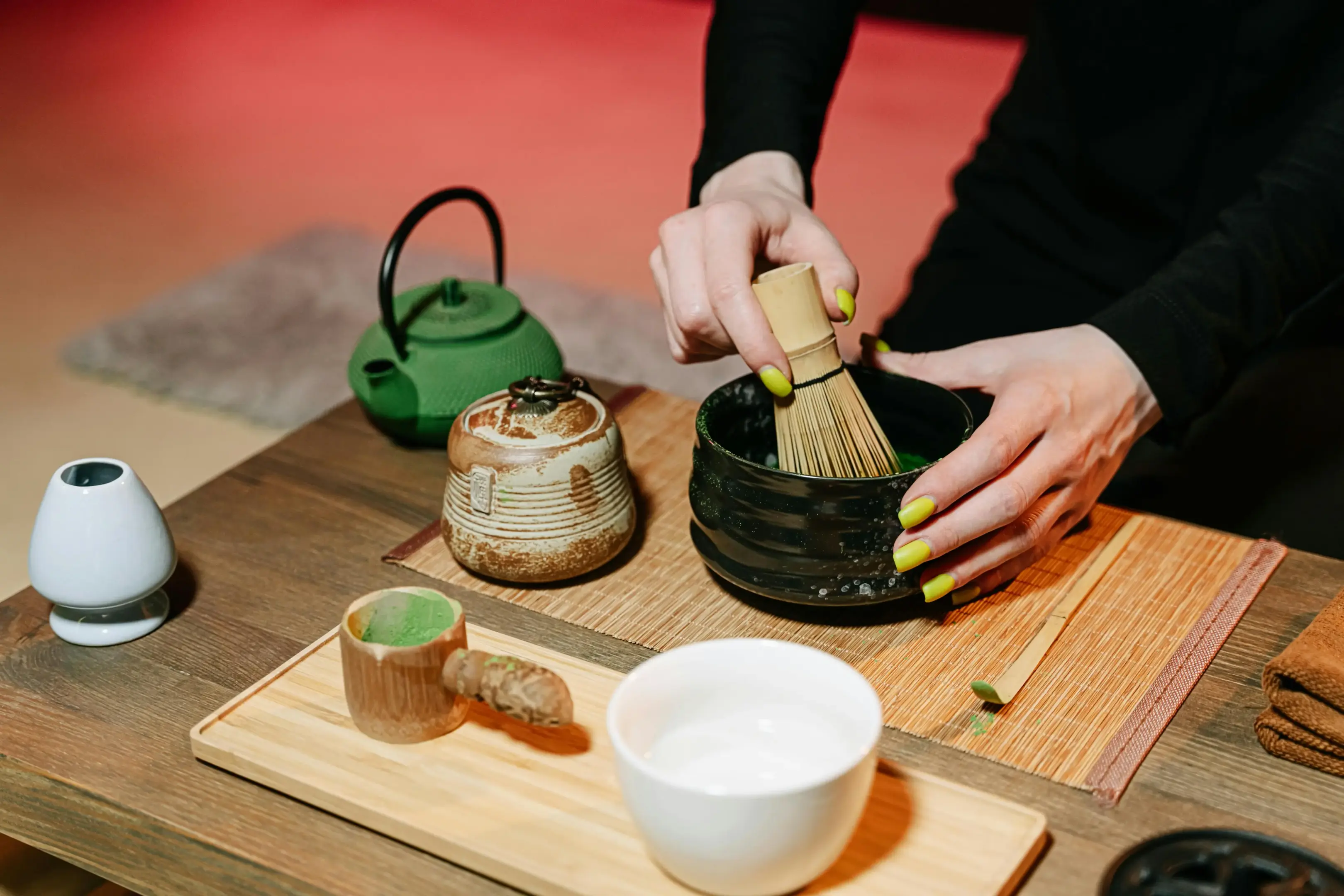 A person with bright yellow nails whisked matcha powder in a black ceramic bowl using a traditional bamboo whisk. On the wooden table, various tea ceremony tools are arranged, including a green cast-iron teapot, a bamboo scoop, and a small container of vibrant green tea powder.
