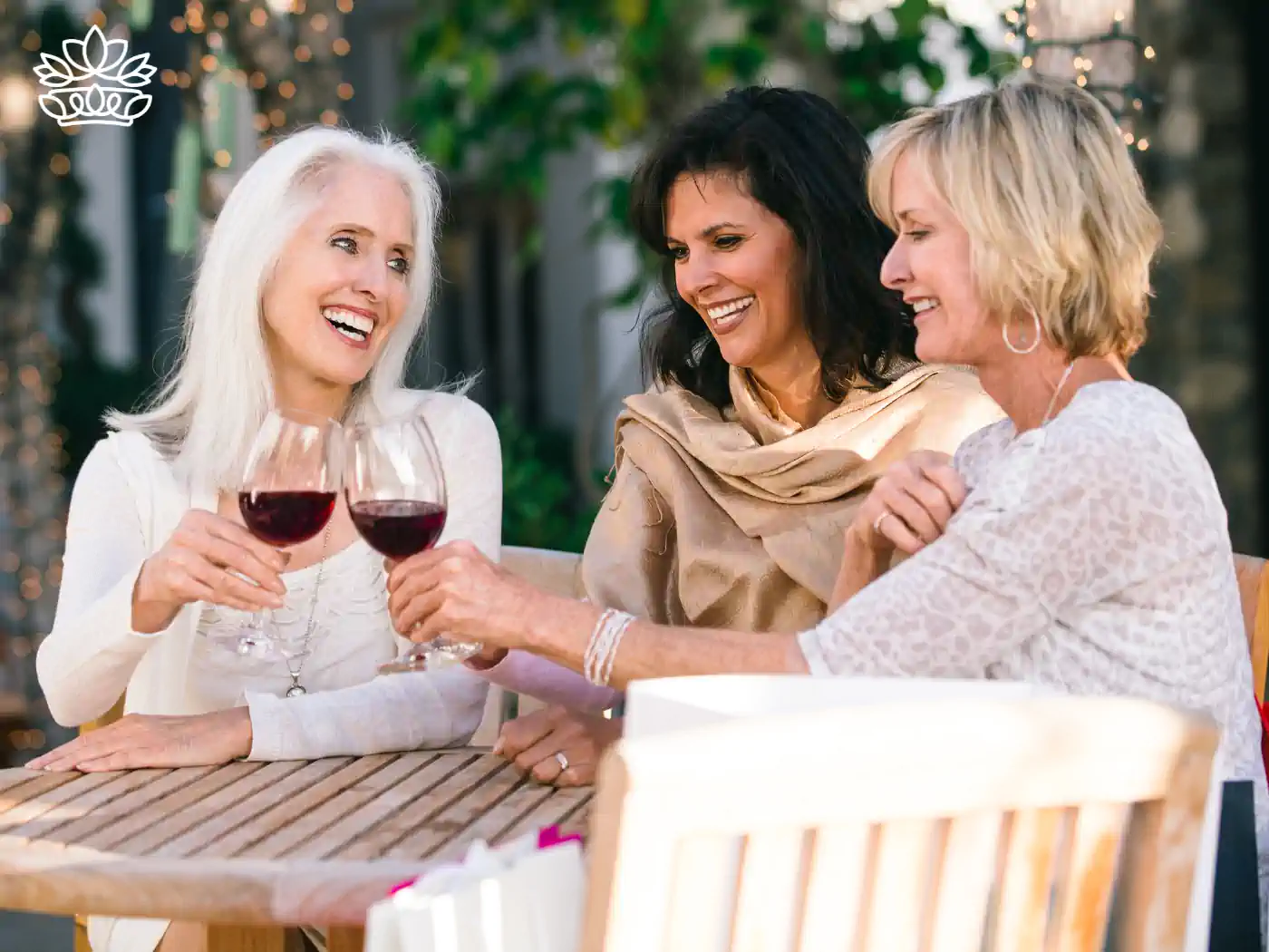 Three women smiling and holding red wine glasses while seated outdoors - Fabulous Flowers and Gifts: Gift Boxes with Champagne and Wine.