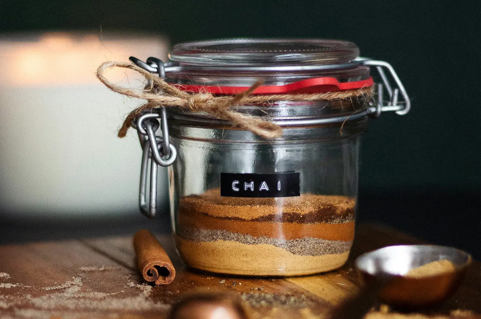 A clear glass jar filled with layered, powdered chai spices is secured with a wire bail lid and rustic twine, labeled with a black sticker reading "CHAI" in white letters, and placed on a wooden surface beside a cinnamon stick and a small metallic scoop.