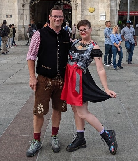 Man in lederhosen in a vest, standing next to a woman in a red and black dirndl. Perfect outfits for Oktoberfest.