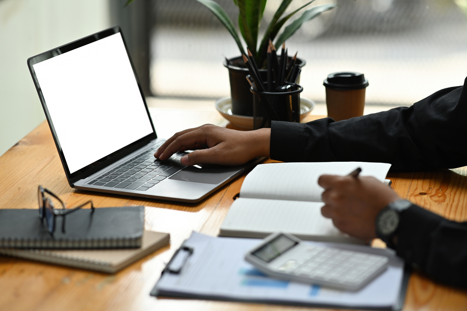 A business professional working on his desk with a notebook, laptop, calculator, and documents.