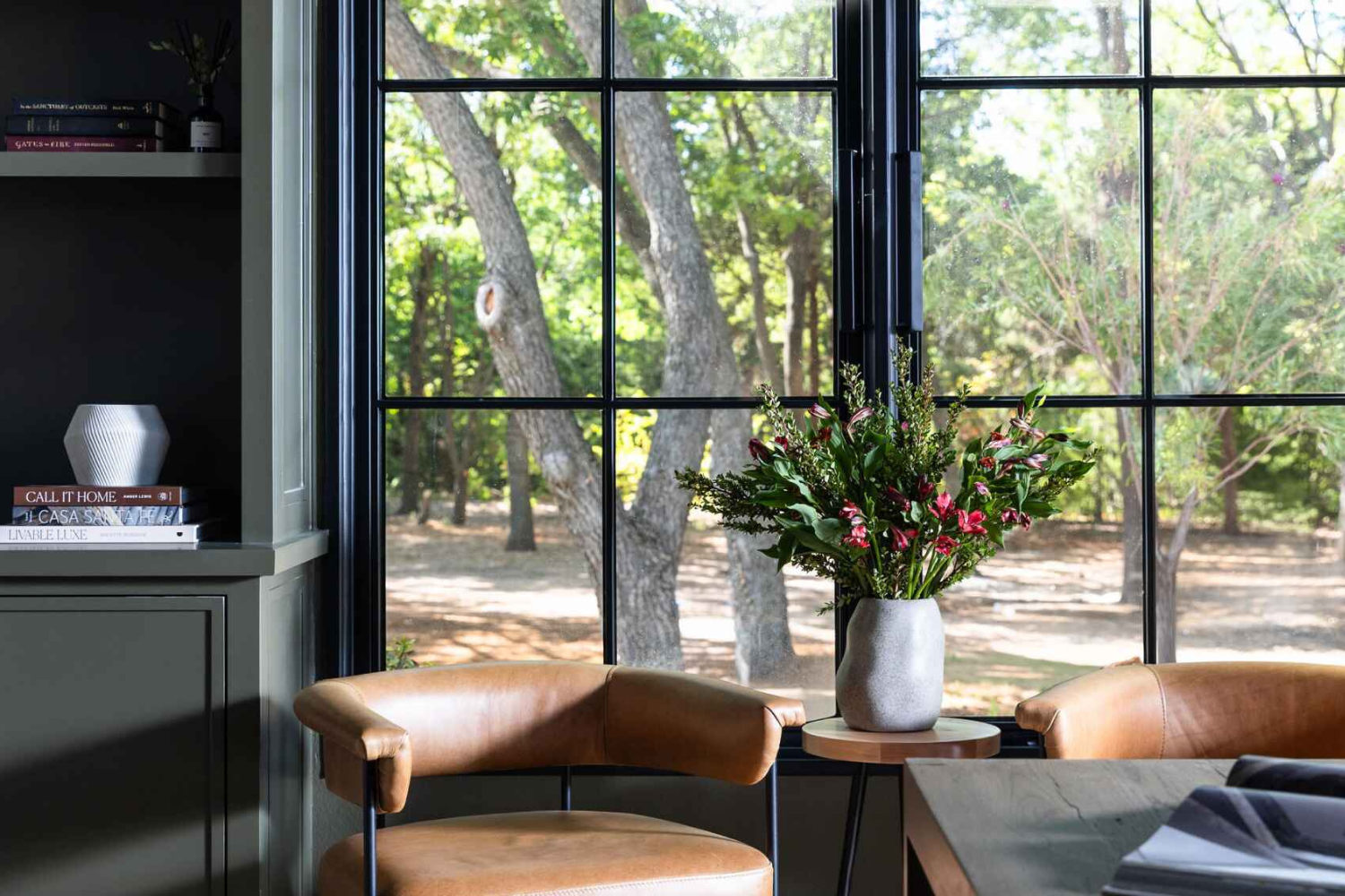 Close-up of a serene office nook with a tan leather armchair beside a vase of fresh greenery, large black steel windows framing a lush outdoor view.