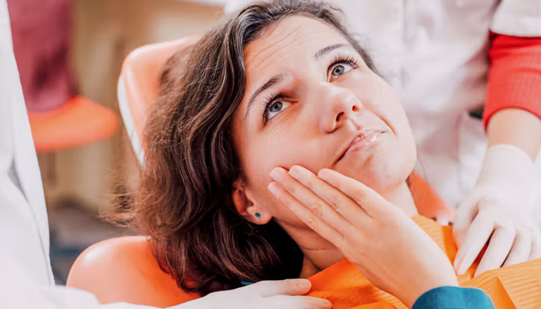 Kvinne sitter i tannlegestol, holder hånden mot kinnet i smerte mens hun ser opp bekymret.