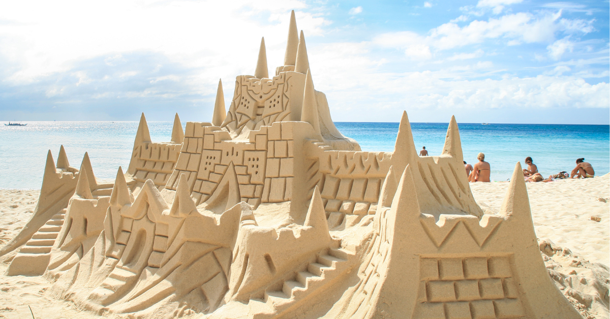 Large, detailed sandcastle sculpture on a sandy beach with people relaxing by the shoreline and turquoise ocean water behind it.