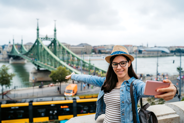 Young woman in a straw hat snapping a selfie in Europe with a bridge behind her.