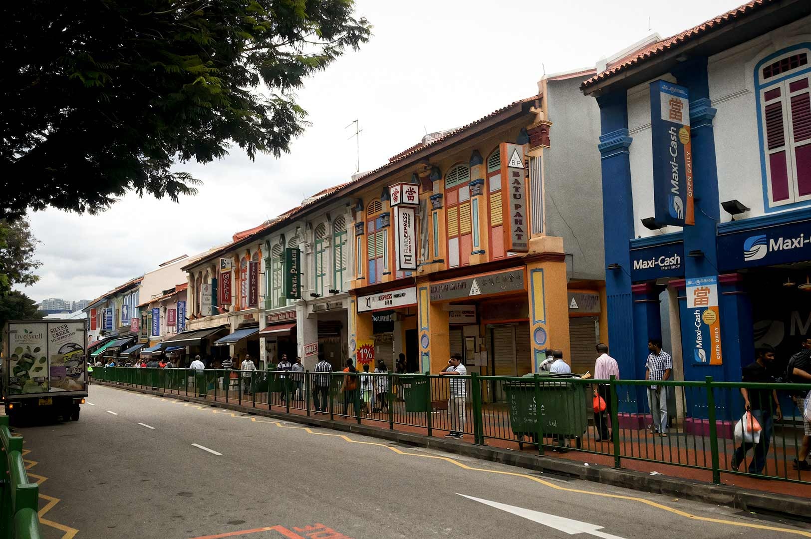 A row of colorful shophouses along Buffalo Road with people walking on the sidewalk beside a fenced street.