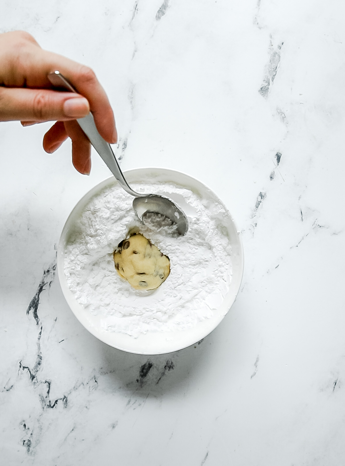 chocolate chip snowball cookie being rolled in a bowl of powdered sugar with a spoon