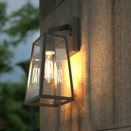 Modern outdoor wall sconce with clear glass panels and black metal frame, mounted on a stone wall and illuminating the surrounding area with a warm light.