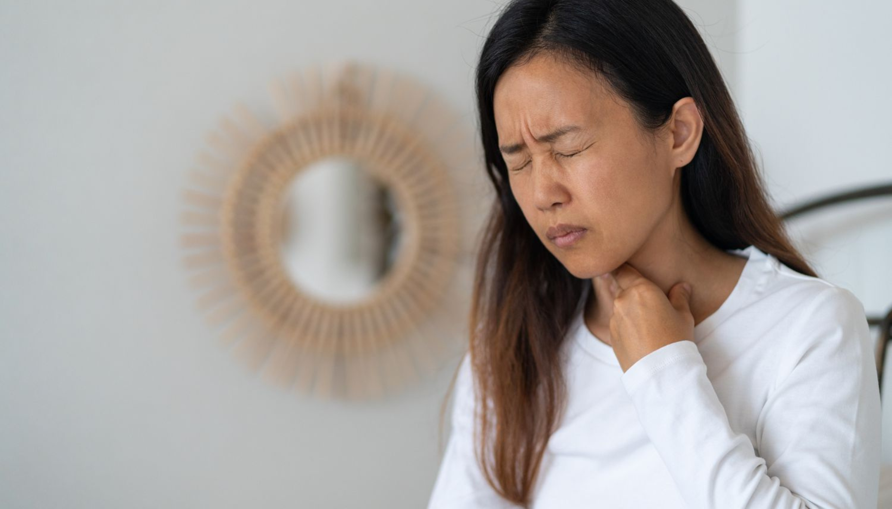 Kvinne i hvit genser holder seg til halsen med et smertefullt uttrykk, tydelig plaget av sår hals eller ubehag.